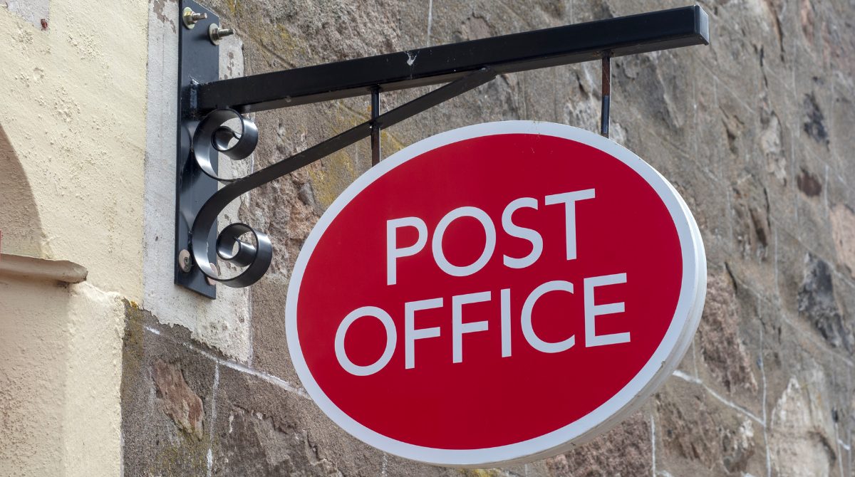 An image of a red and white post office sign, hanging from a black bracket against a sand brick wall.